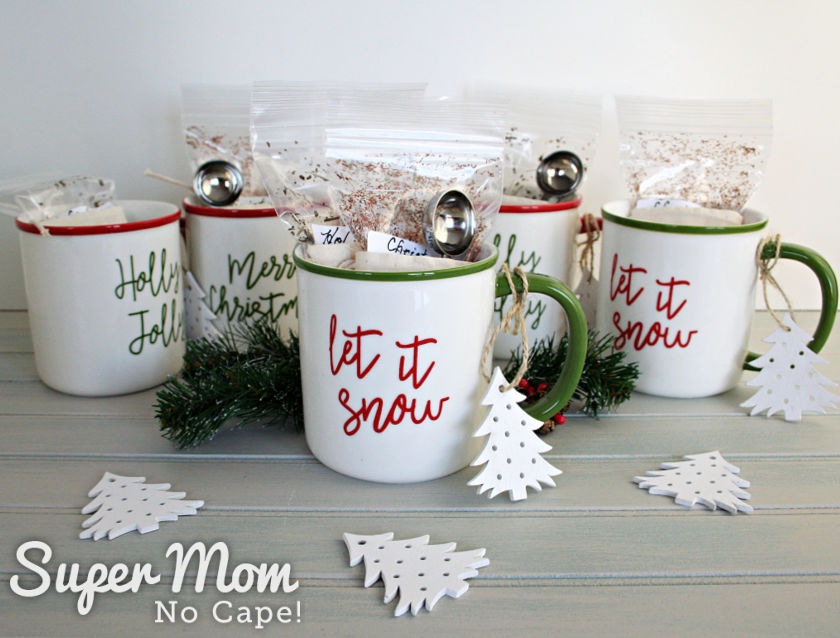 A picture of five tea mugs with bags of loose leaf tea stuffed inside. Each mug has a wooden Christmas Tree-shaped ornament tied to the handle.